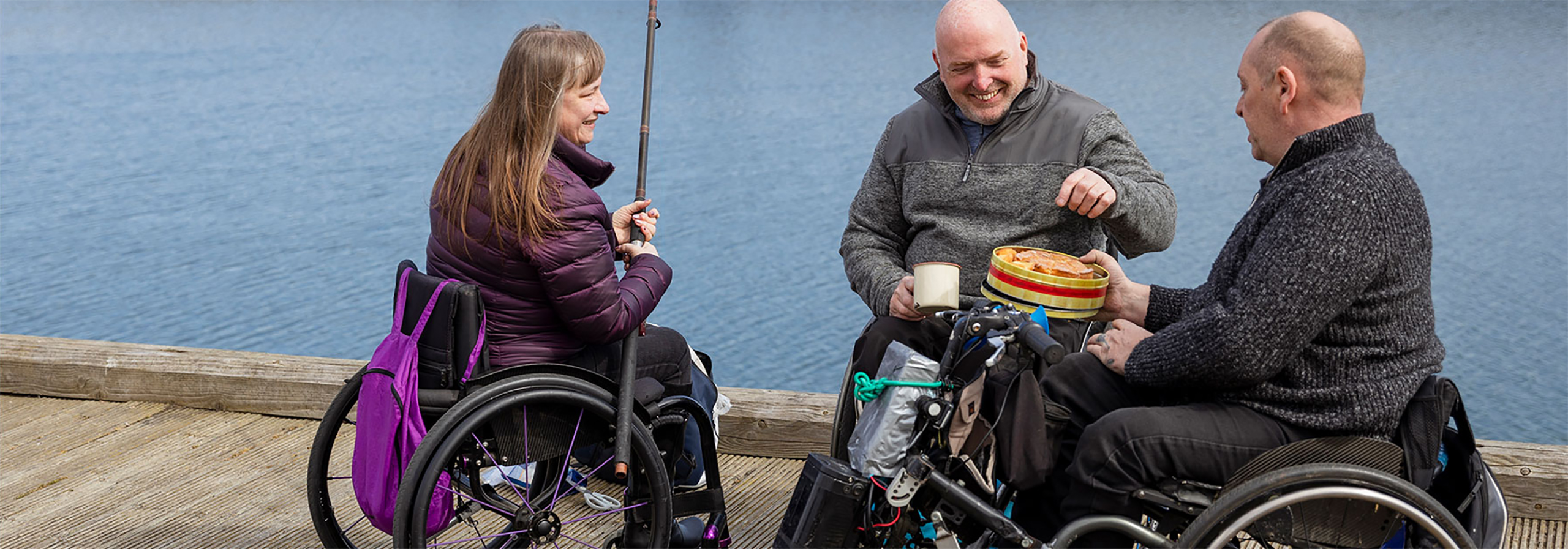 three people in a wheelchairs sitting on a dock, fishing, eating and smiling