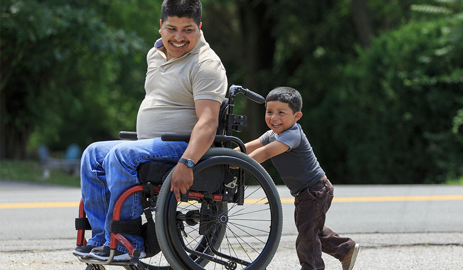 Young father in a wheelchair being pushed by his son.