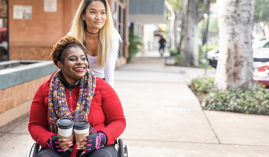 Young African American female wheelchair-user rolling on the sidewalk with her friend.