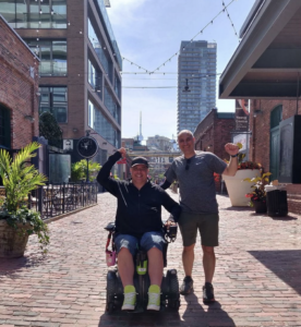 Man in wheelchair and man standing; waving at camera; hanging out outdoors.
