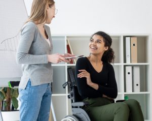 A young woman in a wheelchair is smiling up at a female friend.