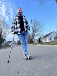 Man with cane in standing outside on road, smiling.